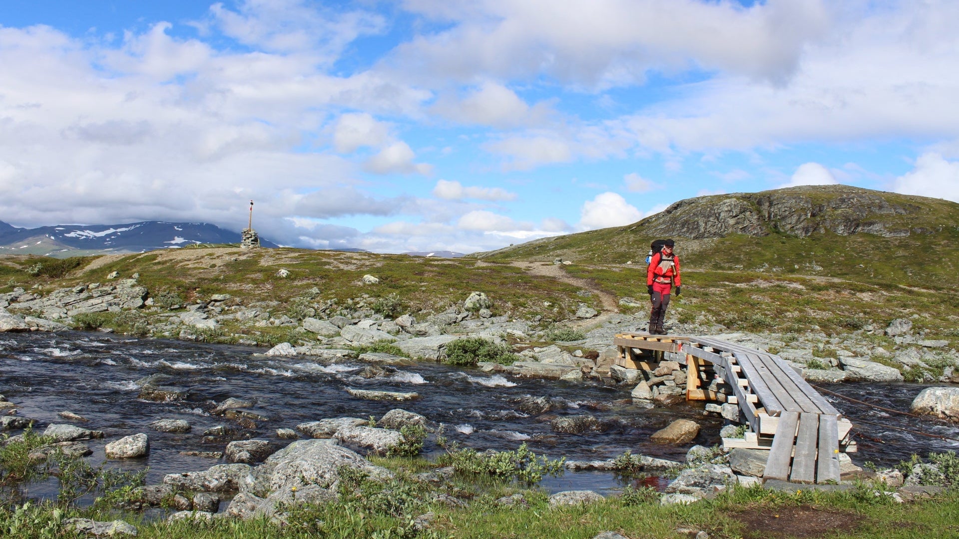 Person standing on a wooden bridge over a stream in a mountainous landscape with a clear blue sky.