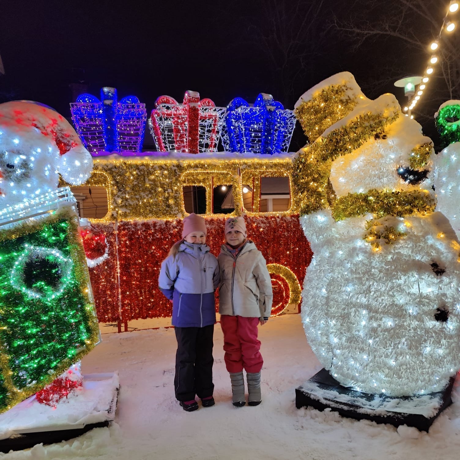 Two children standing in front of a decorated house with lights and a large snowman at night.