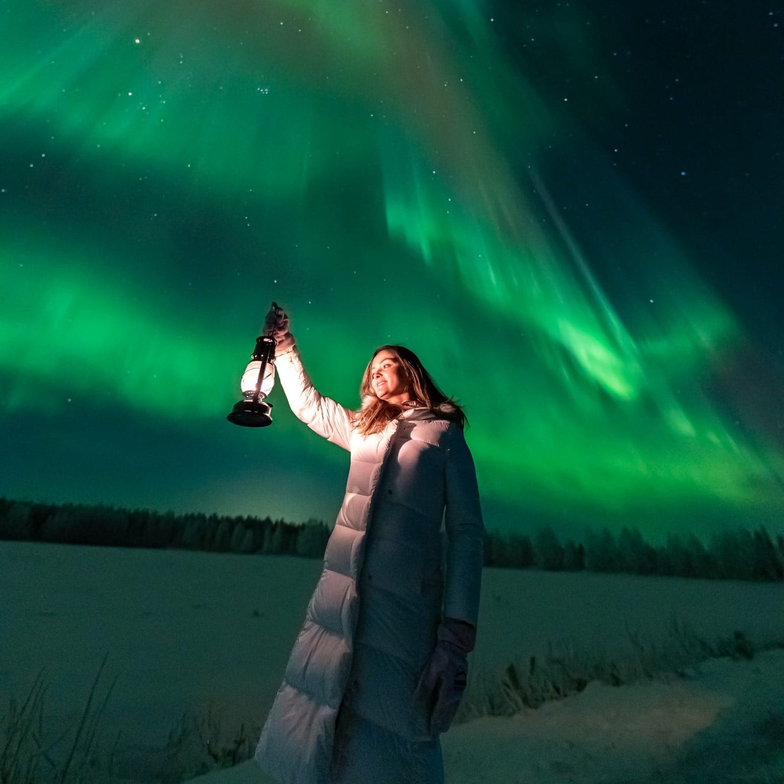 Woman holding lantern under vibrant Northern Lights on Aurora Borealis Hunting Pro Guaranteed Tour by Book Lapland
