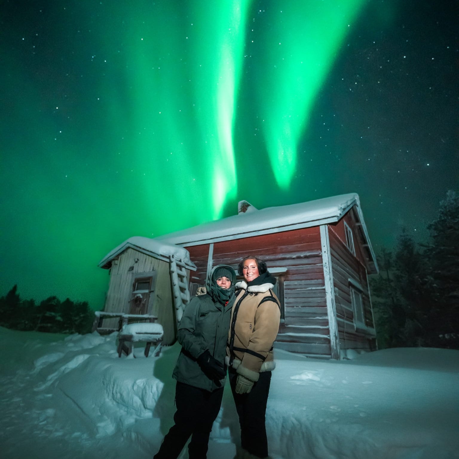 Aurora Borealis Hunting Pro Guaranteed Tour shows two people near a cabin under bright Northern Lights by Book Lapland