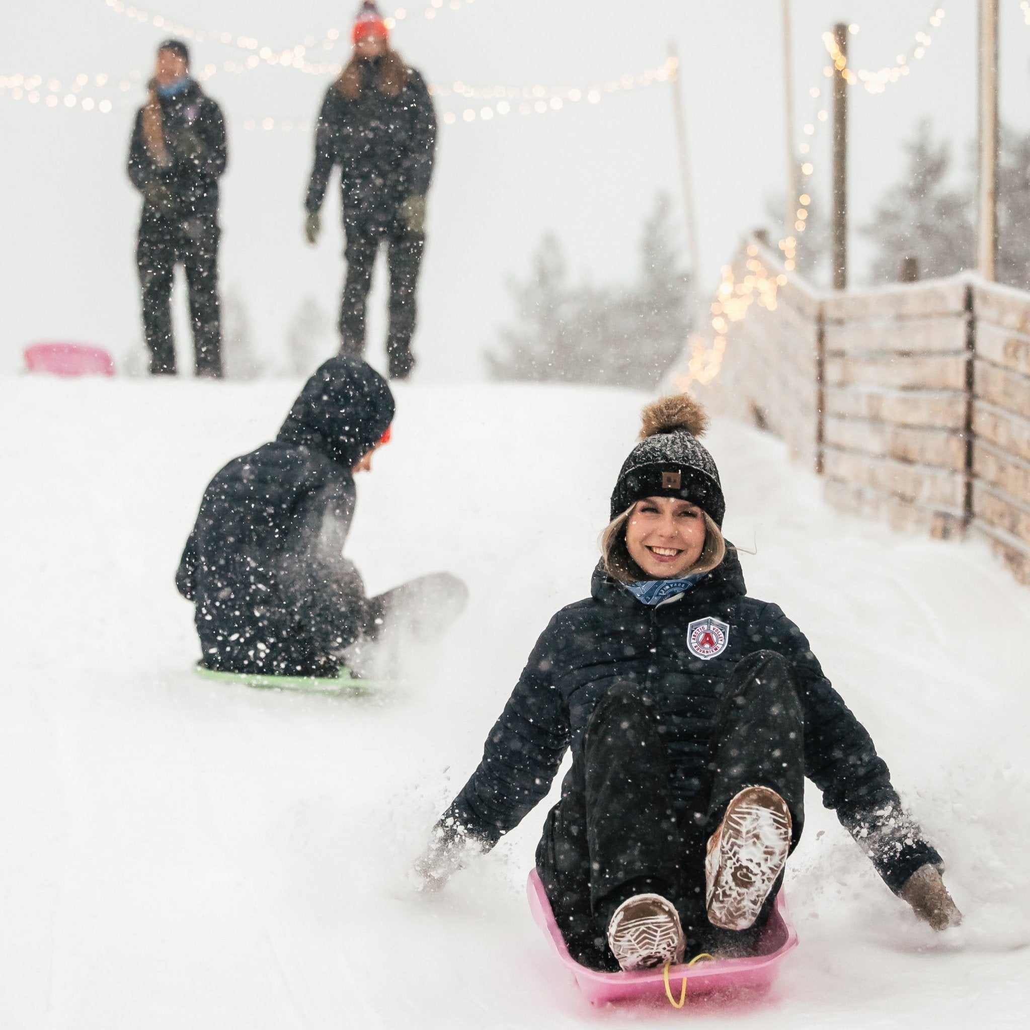 Two people sledding down a snowy hill with a festive atmosphere.
