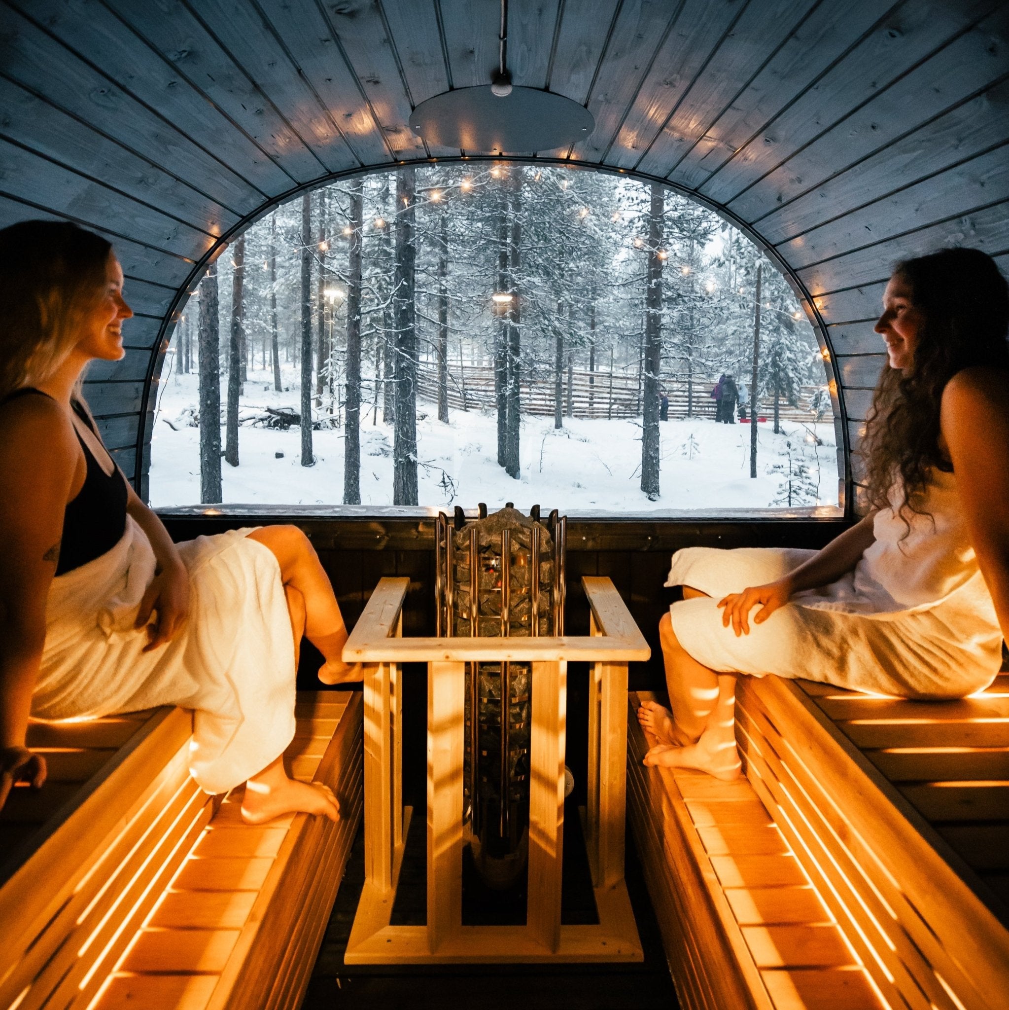 Two people sitting in a wooden sauna with a snowy forest view outside.