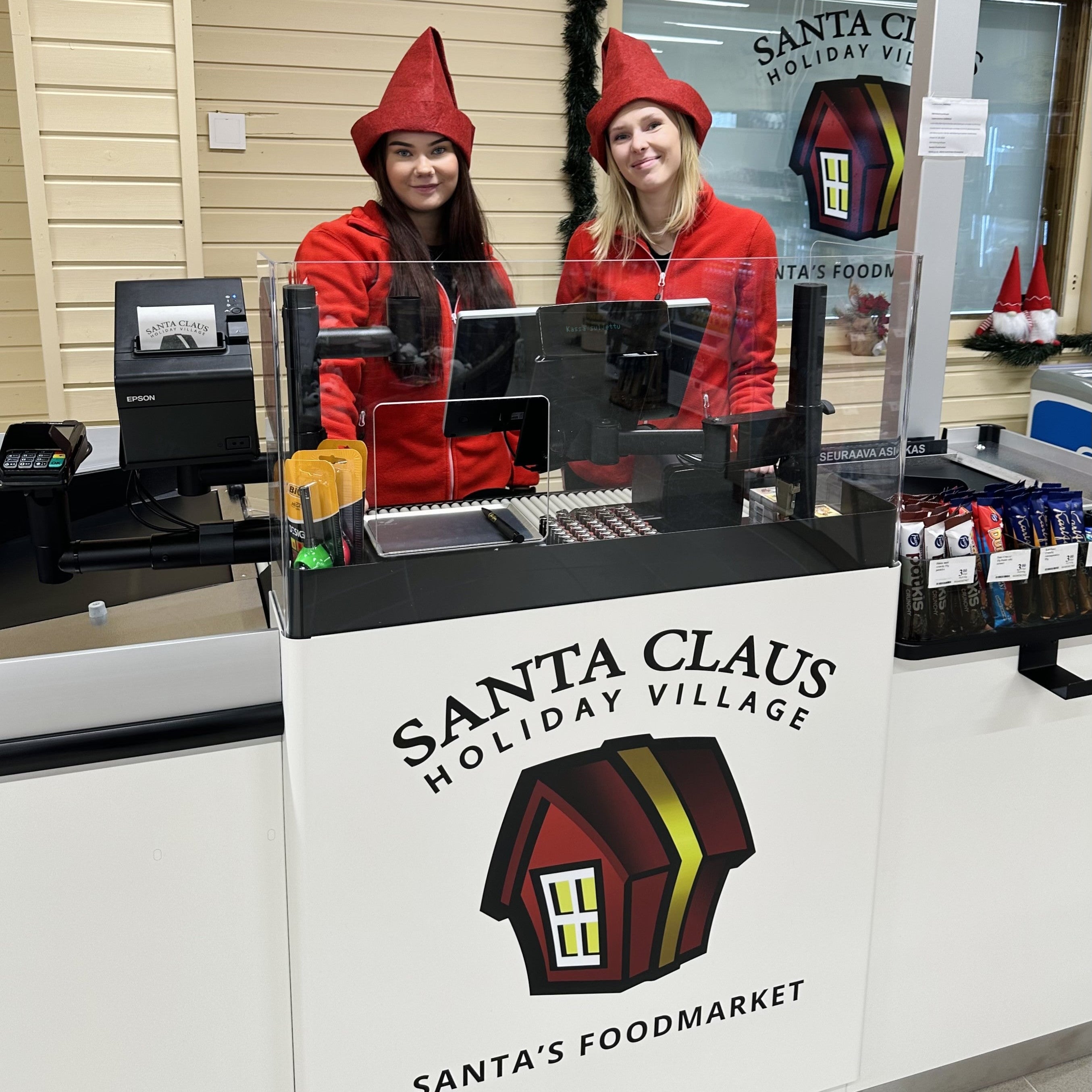 Two people in red outfits behind a counter at Santa Claus Holiday Village with a sign in the background.
