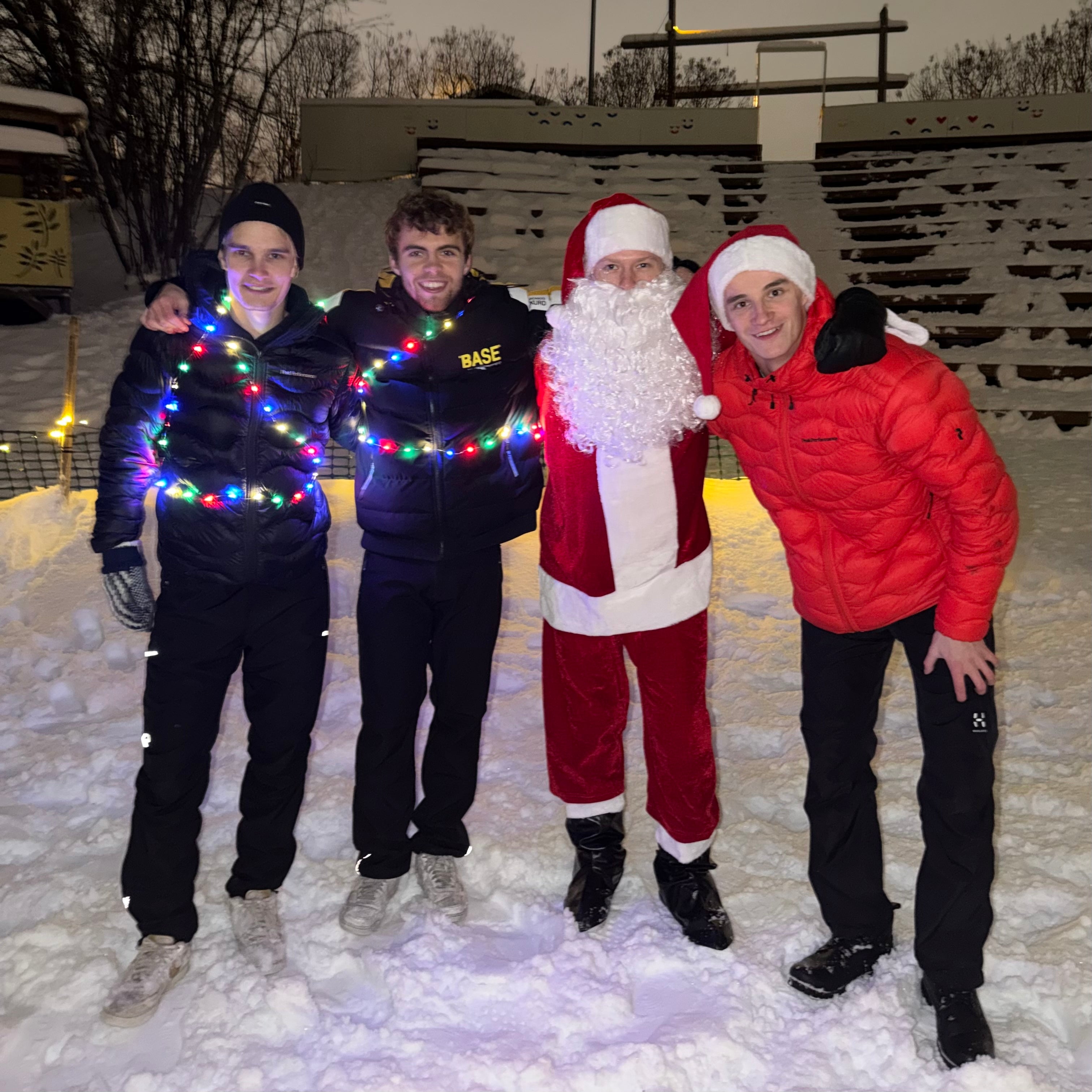Four people, including a person in a Santa Claus costume, standing in the snow with Christmas lights.