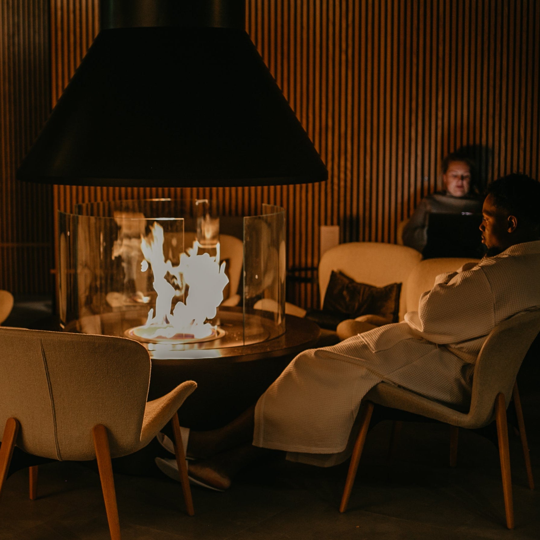 Person relaxing by indoor firepit wearing bathrobe during Arctic spa experience with bathrobe dinner Invisible forest lodge