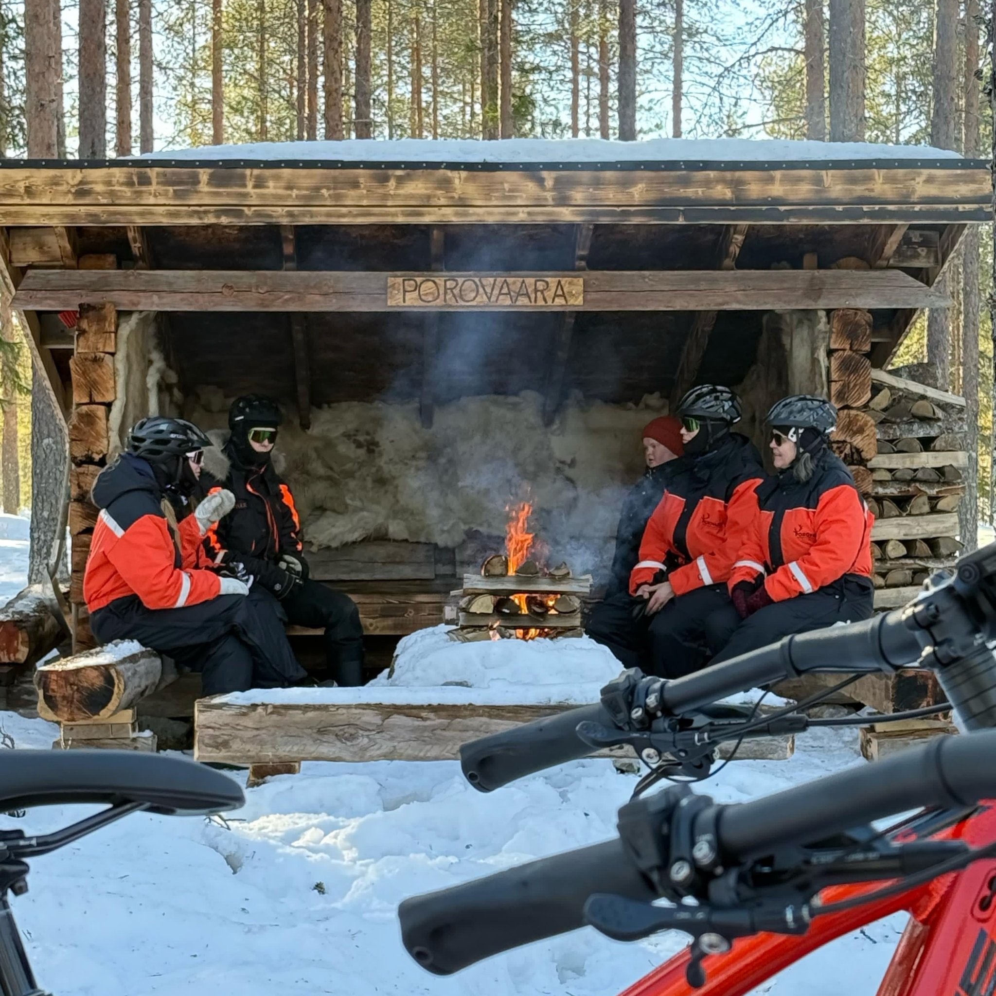 Group wearing helmets resting by a fire in a snowy shelter during Electric Fat Bike Adventure at a Reindeer