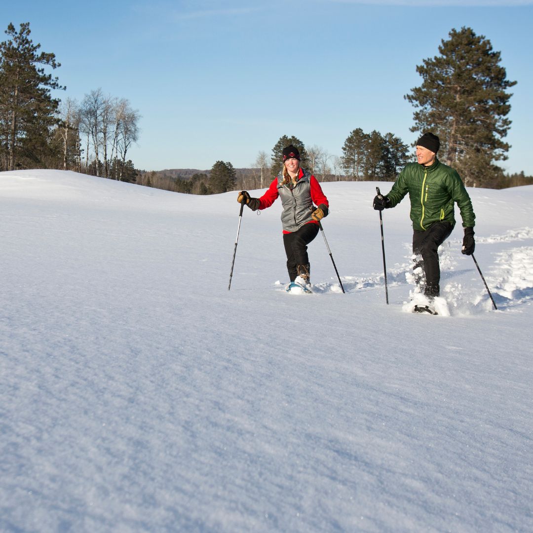 Two people cross-country skiing in a snowy winter landscape with trees in the background.